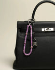 Leah Bag Charm attached to a black handbag – The lavender quartz charm clipped onto a black designer handbag, showcasing its elegant and energy-infused accent.