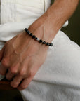 A man wearing the David Bracelet in Silver, featuring 6mm Onyx and Black Labradorite Quartz beads with a black CZ diamond evil eye charm.