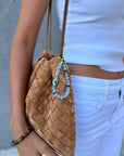 A tan woven suede bag with thin brown straps, styled with a blue Mini Minou charm made of aquamarine quartz beads and gold accents, clipped to the bag. The model wears a white crop tank, white jeans, and a brown beaded bracelet.