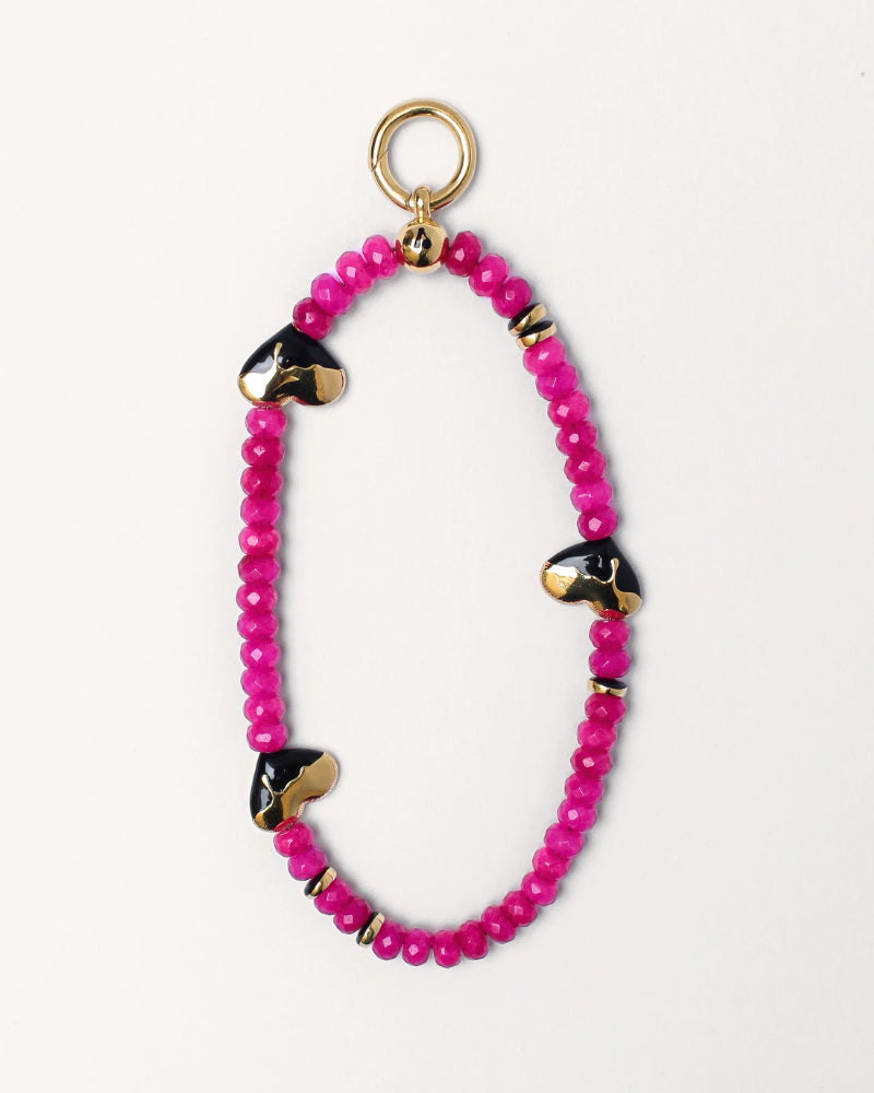 The Aria wristlet in pink quartz featuring vibrant pink beads, gold-plated heart-shaped charms with black detailing, and a gold ring attachment, combining playful elegance and functional design.