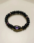 Aya bracelet with onyx quartz beads, accented with purple and gold dividers, featuring an evil eye charm with a purple CZ stone."