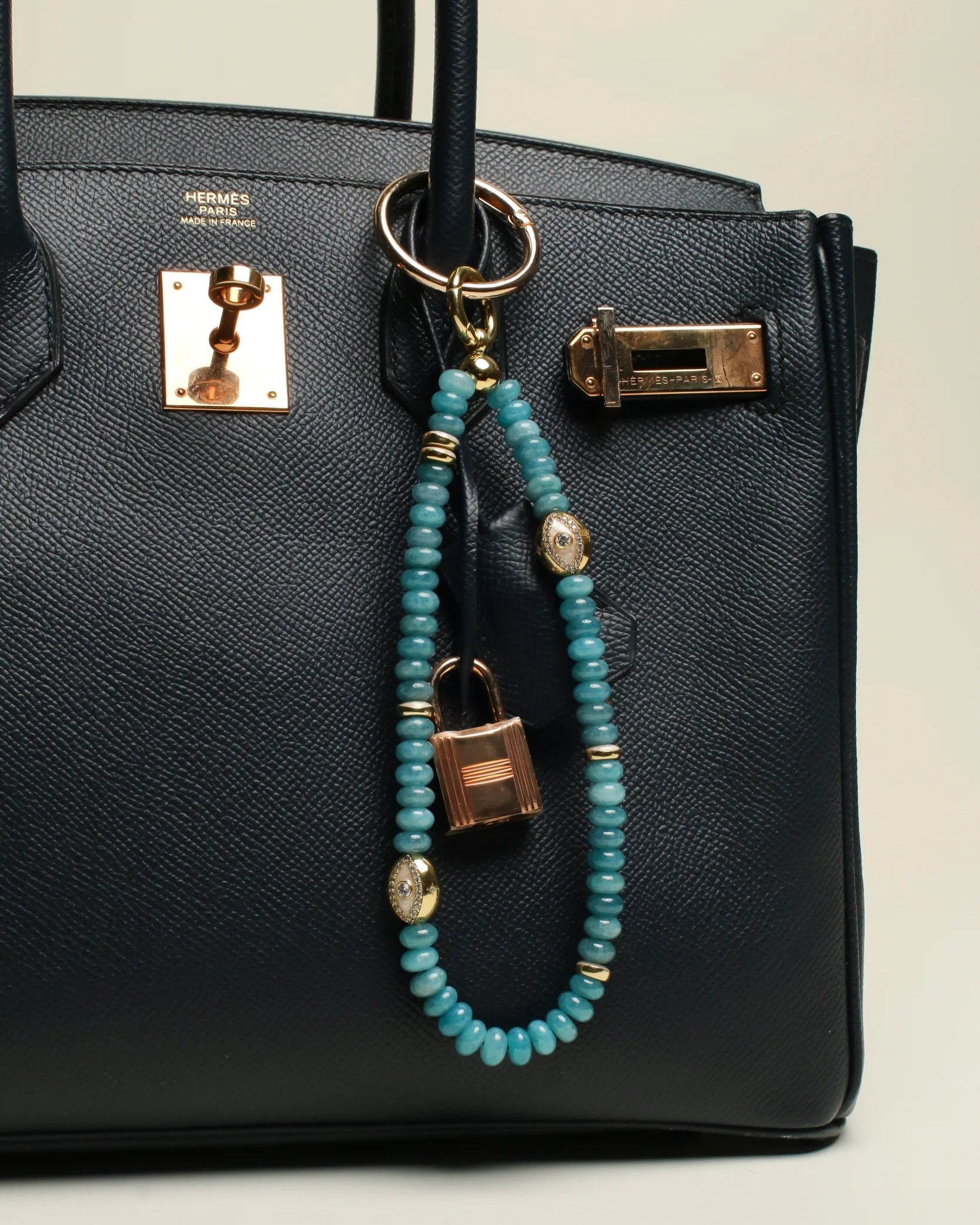 A black designer handbag with The Leah Bag Charm in Apatite attached to the handle. The charm features semi-precious apatite quartz beads, gold-plated Evil Eye charms, and luxe dividers, adding a refined and stylish touch.