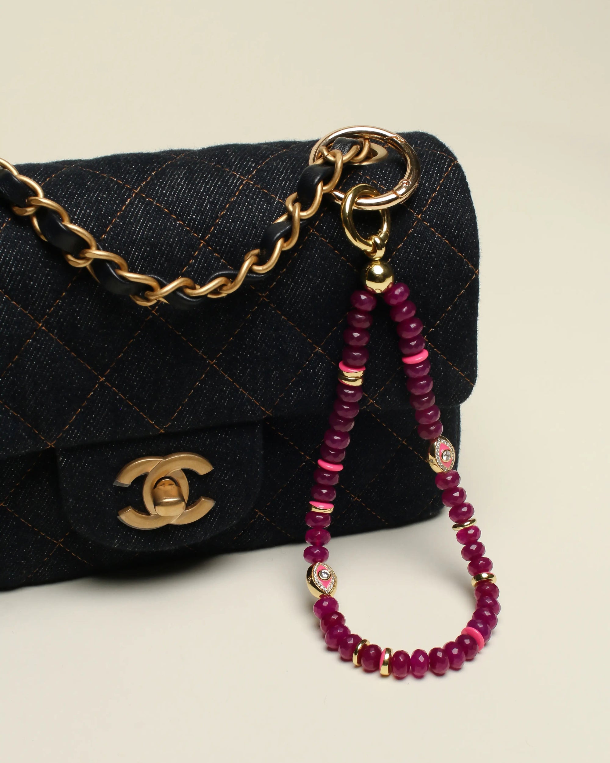 Leah Bag Charm in Amethyst attached to a quilted denim Chanel bag – A faceted amethyst quartz bag charm with 14k gold-plated accents and Evil Eye charms clipped onto a luxurious quilted denim Chanel handbag, highlighting its elegant and protective design.