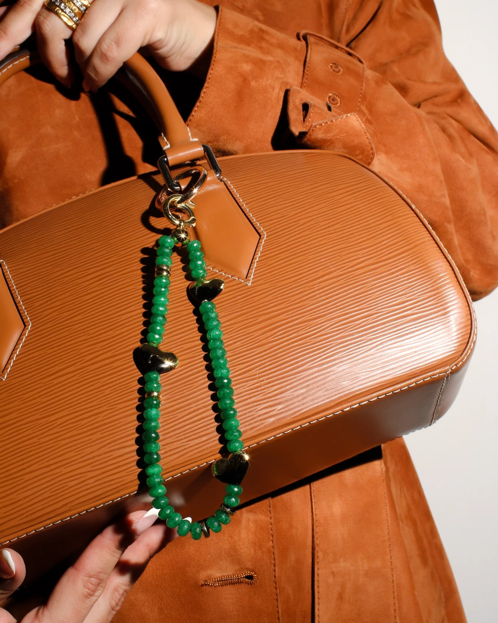 A model carries a brown leather handbag adorned with The Aria Bag Charm in Emerald, highlighting its bold emerald quartz beads and gold-plated heart charms for a luxurious, polished look.