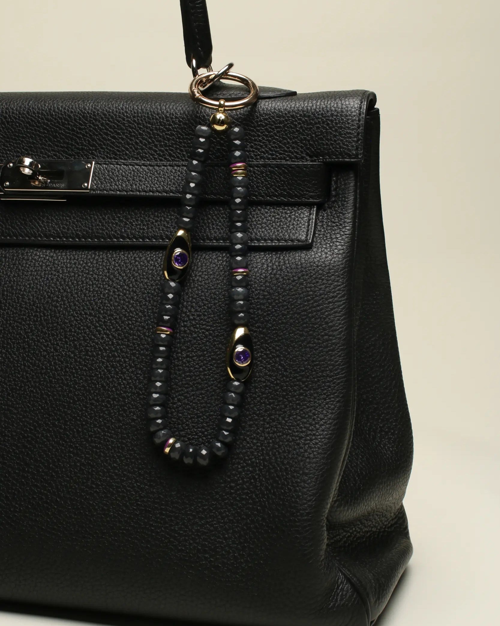 A black designer handbag with The Aya Bag Charm in Onyx attached to the handle. The charm features semi-precious onyx beads, gold-plated Evil Eye charms with amethyst stones, and luxe dividers, adding a bold and protective touch.