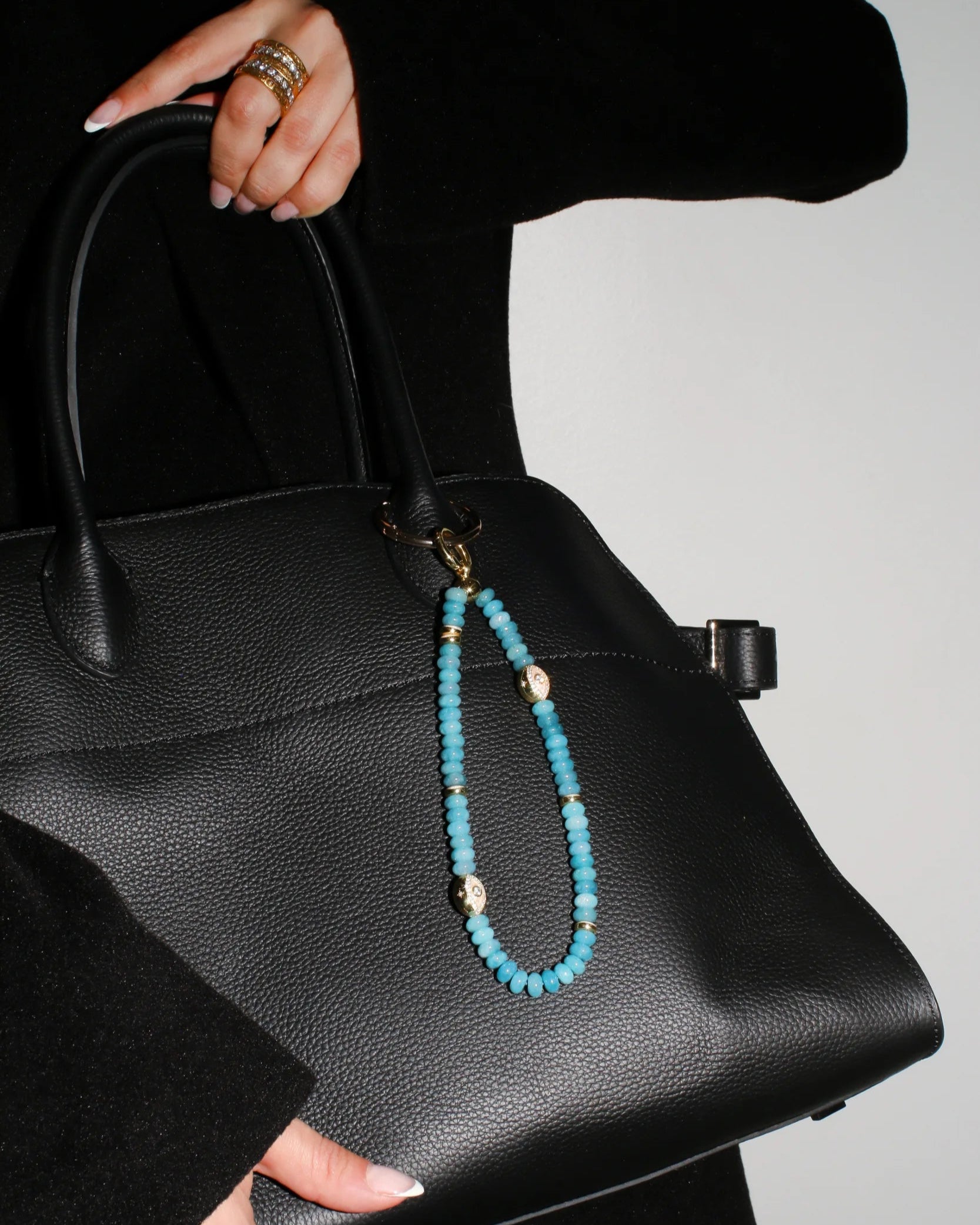 A model carries a black handbag adorned with The Leah Bag Charm in Apatite, showcasing its elegant design and protective symbolism through gold-plated Evil Eye charms and rich apatite quartz beads.