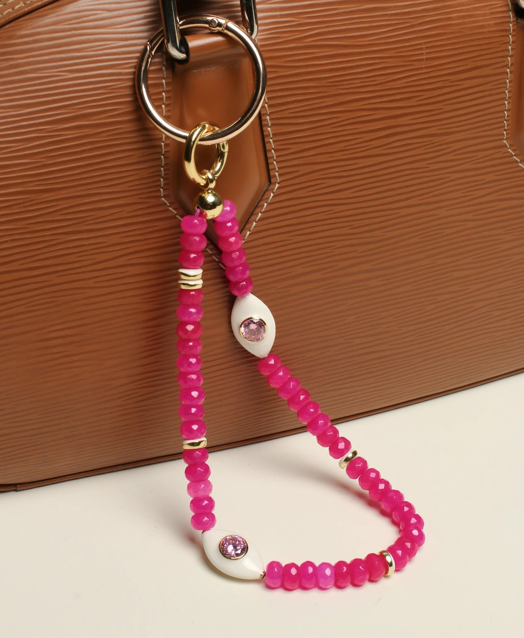  A structured brown handbag with The Mano Bag Charm in Pink attached to the handle. The charm features semi-precious pink quartz beads, eye-shaped charms with CZ diamonds, and luxe gold-plated accents, adding a bold and stylish touch.