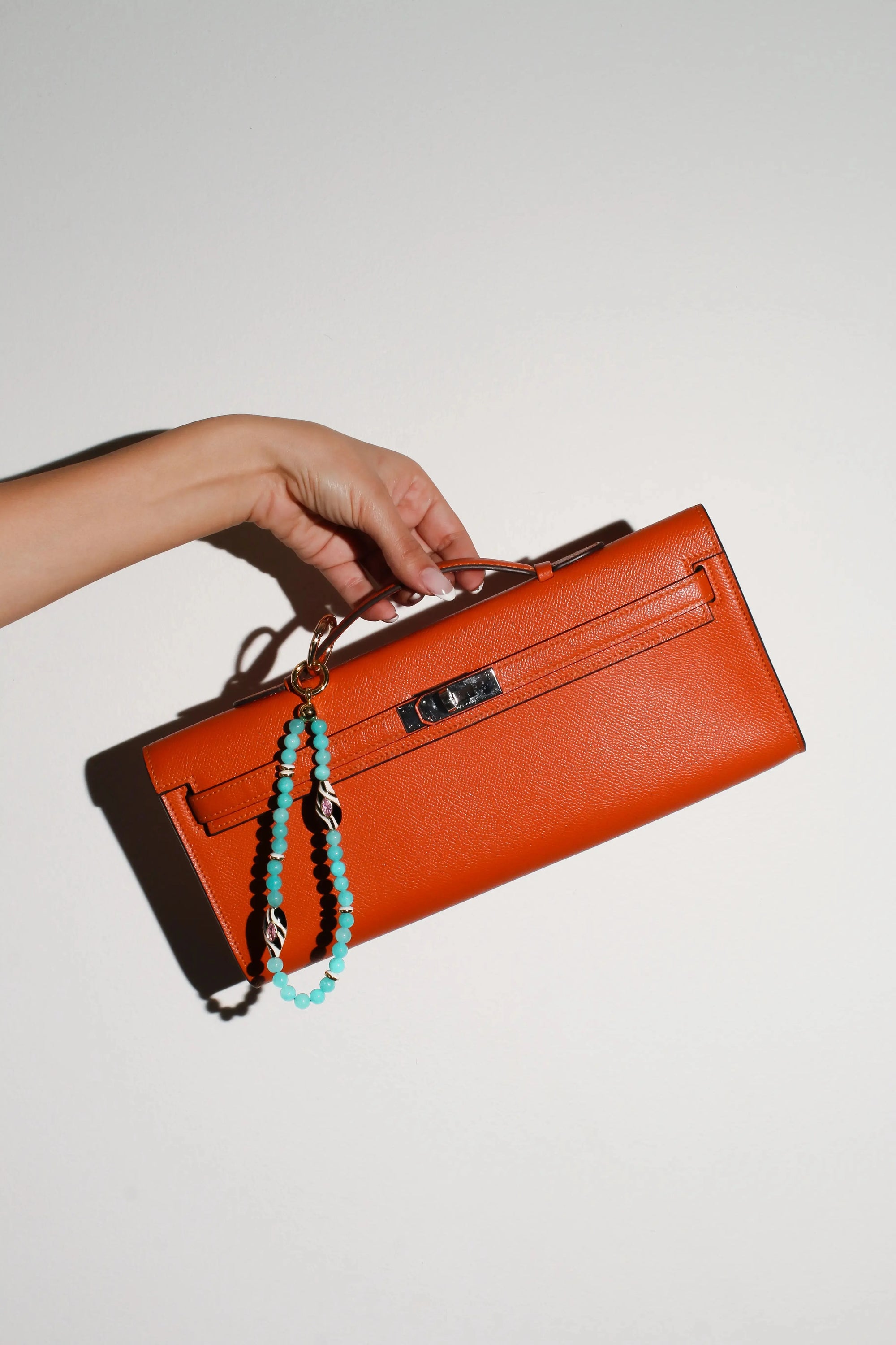 A model holds an orange handbag with The Minou Bag Charm in Aqua attached, featuring semi-precious aqua quartz beads and a gold-plated zebra-striped Evil Eye charm for a bold, protective accent.