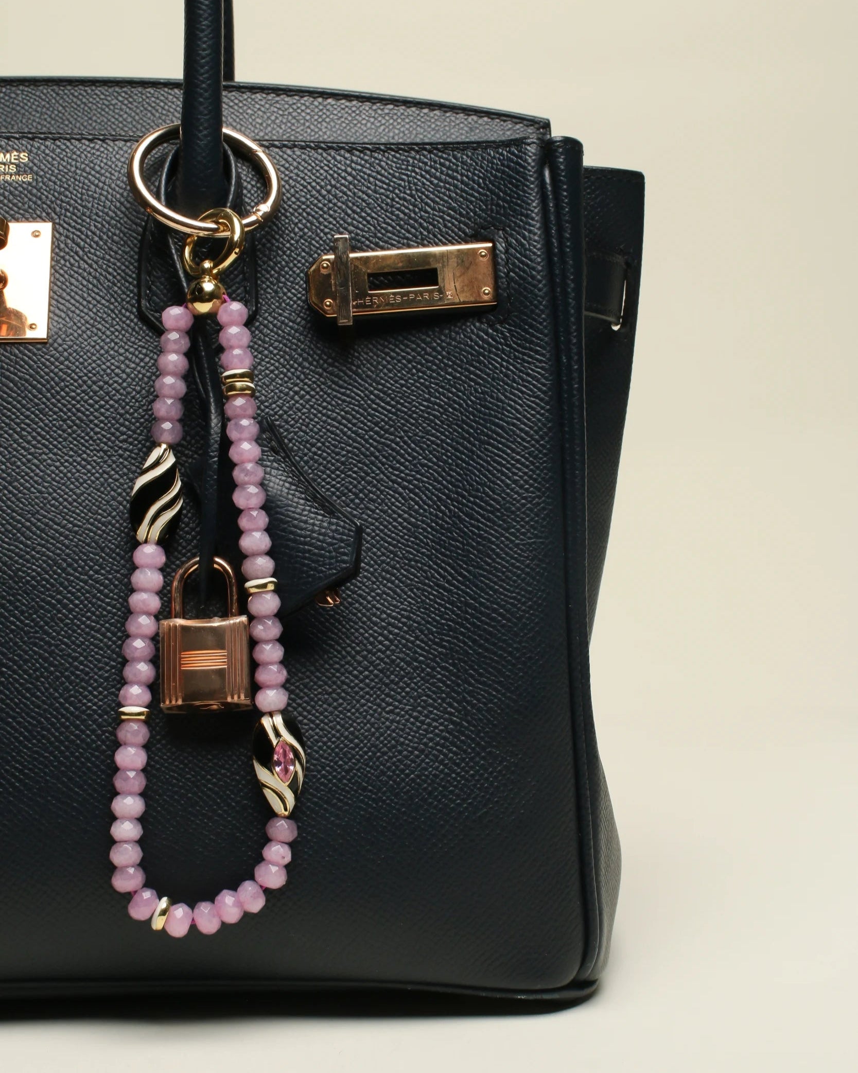 A black designer handbag with The Minou Bag Charm in Lavender attached to the handle. The charm features semi-precious lavender quartz beads, a zebra-striped Evil Eye charm, and gold-plated details, adding an elegant and protective touch.