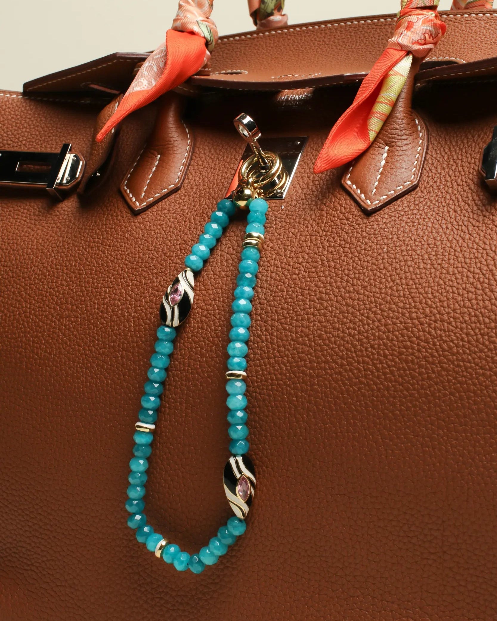 A brown designer handbag with The Minou Bag Charm in Turquoise attached to the handle. The charm features semi-precious turquoise quartz beads, gold-plated zebra-striped Evil Eye charms, and luxe dividers, adding a bold and protective touch.