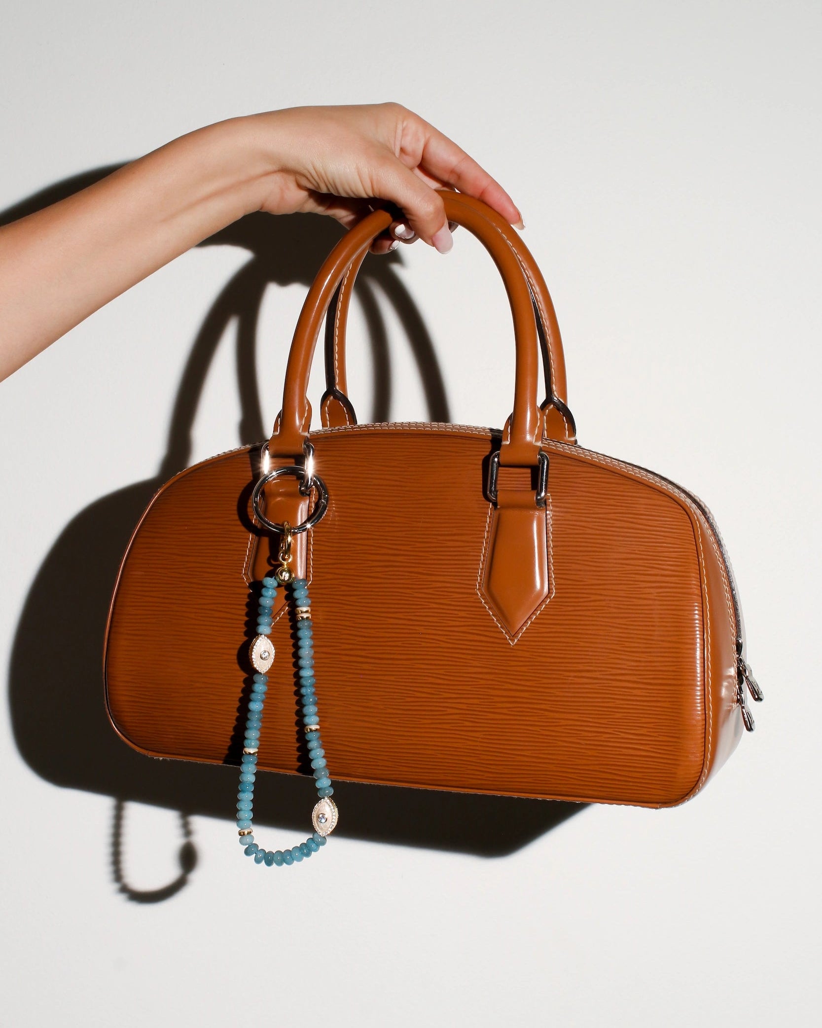 A brown leather handbag with The Stella Bag Charm in Apatite attached to the handle. The charm features semi-precious apatite quartz beads, a gold-plated Evil Eye charm, and zirconia diamonds, adding a chic and elegant touch.