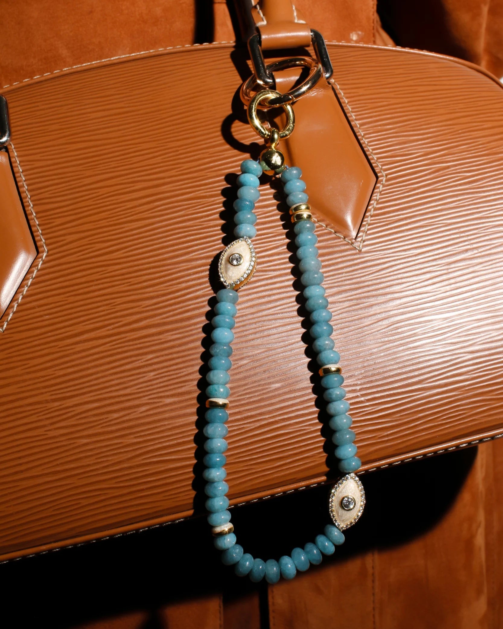 The Stella Bag Charm in Apatite elegantly draped over a handbag, showcasing its luxurious design with gold-plated accents, apatite quartz beads, and an Evil Eye charm for a stylish and protective touch.