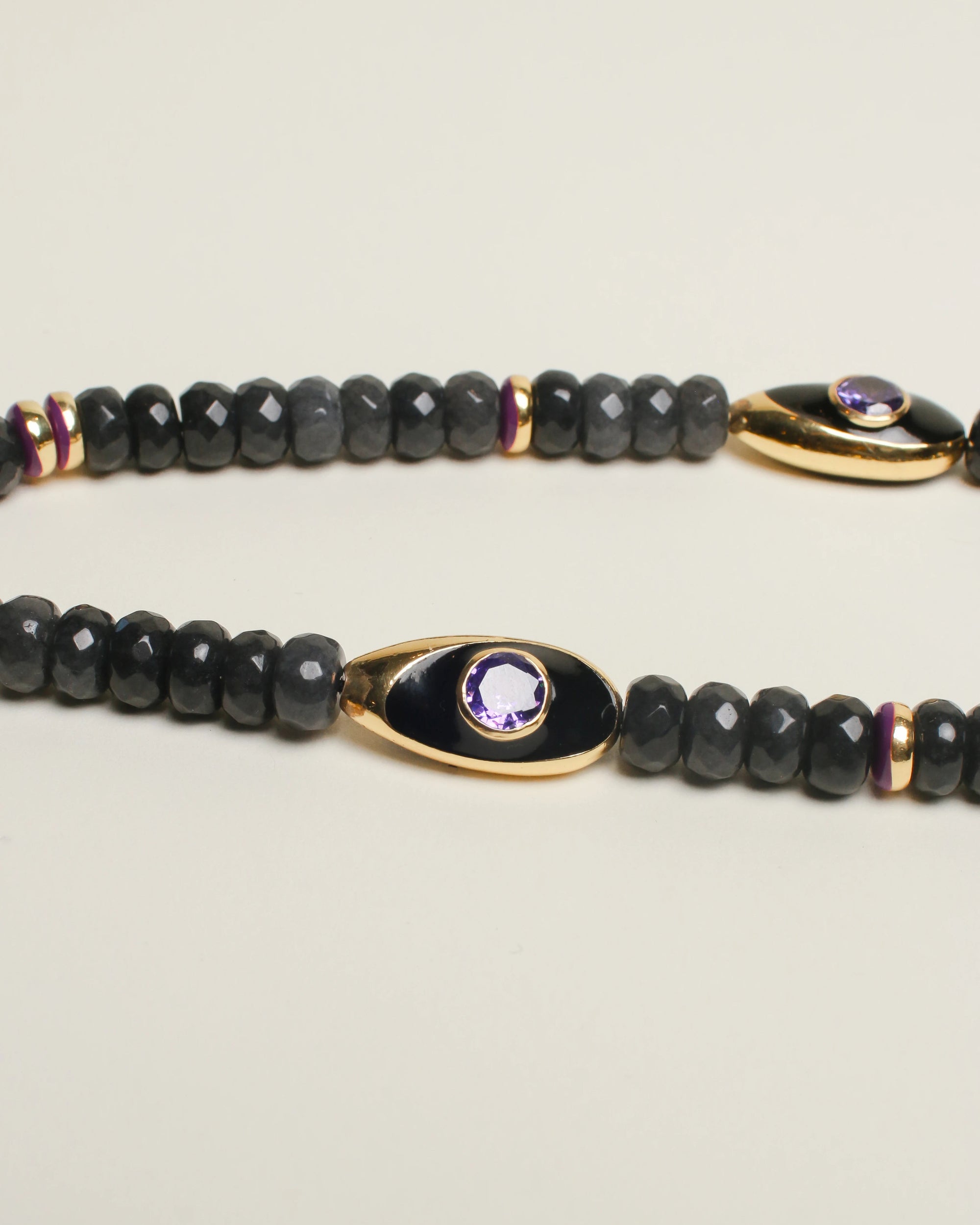 A close-up of The Aya Bag Charm in Onyx, highlighting the intricate design of the gold-plated Evil Eye charms, shimmering amethyst stones, and polished onyx beads.