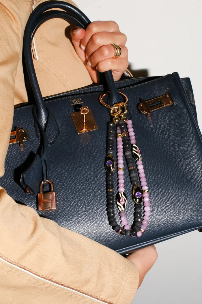A model carries a black handbag featuring The Minou Bag Charm in Lavender, showcasing its stylish, protective design with lavender quartz beads, gold-plated details, and a bold Evil Eye charm.