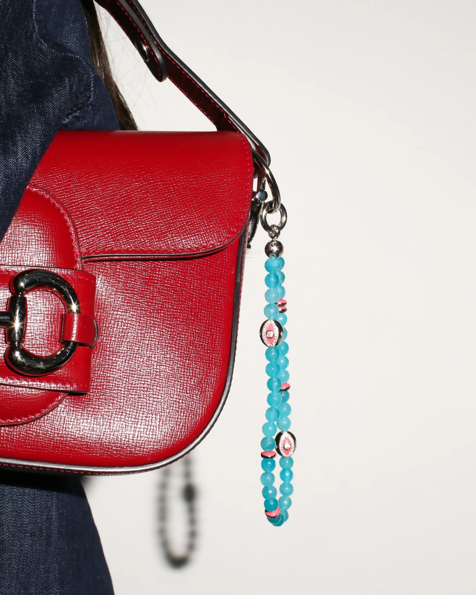 A red handbag with The Leah Bag Charm in Turquoise attached to the strap. The charm features semi-precious turquoise quartz beads, rhodium-plated silver Evil Eye charms, and pink dividers, adding a vibrant and protective touch.