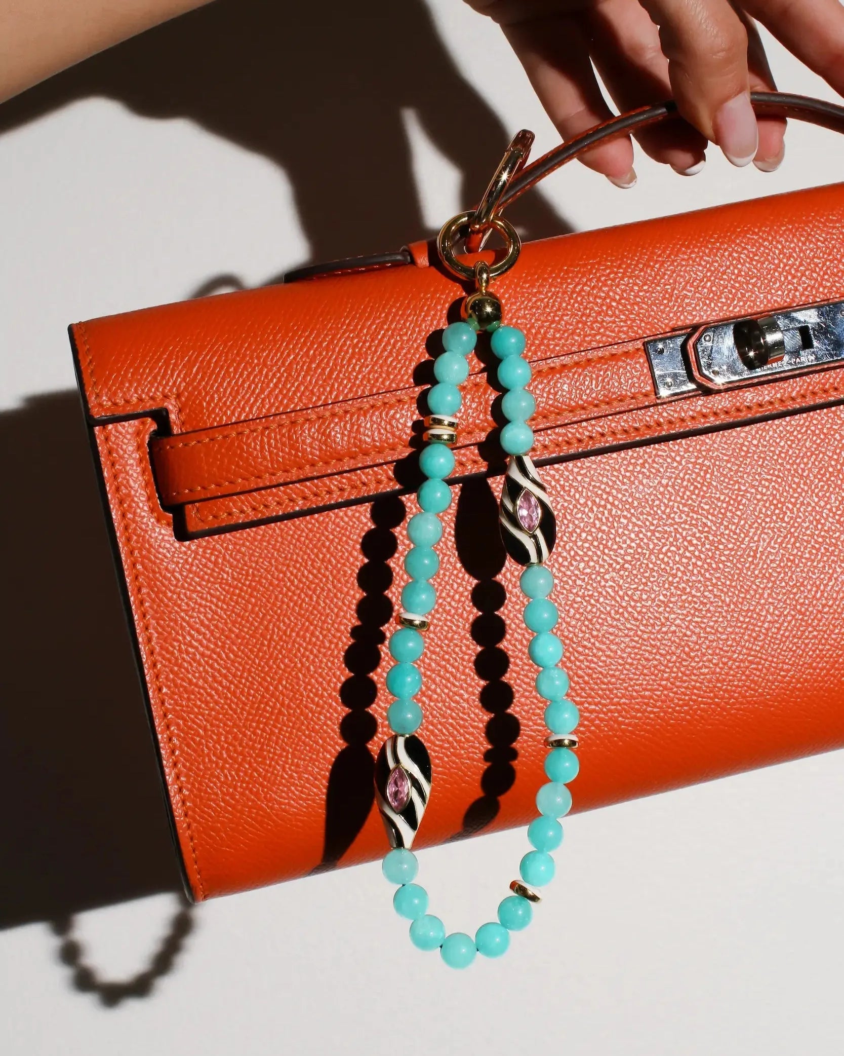 A model carries an orange handbag featuring The Minou Bag Charm in Aqua, showcasing its elegant and protective design with aqua quartz beads and gold-plated details.