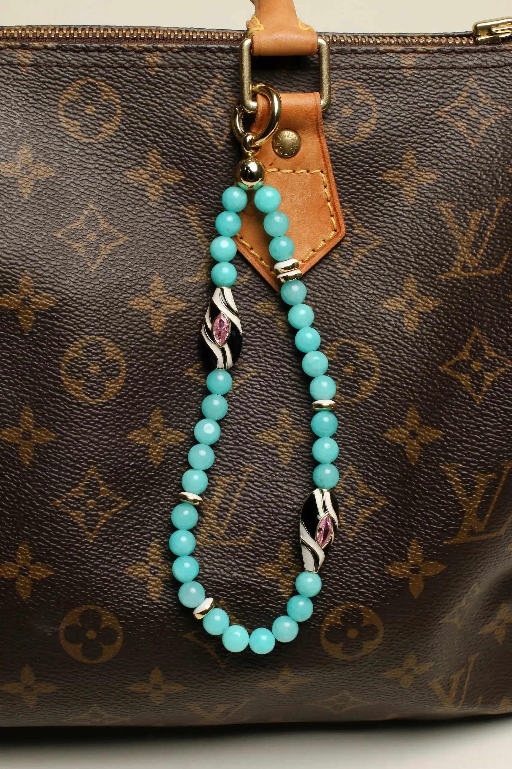 A brown designer handbag adorned with The Minou Bag Charm in Aqua, highlighting its vibrant aqua quartz beads and gold-plated Evil Eye charm for an elegant touch.