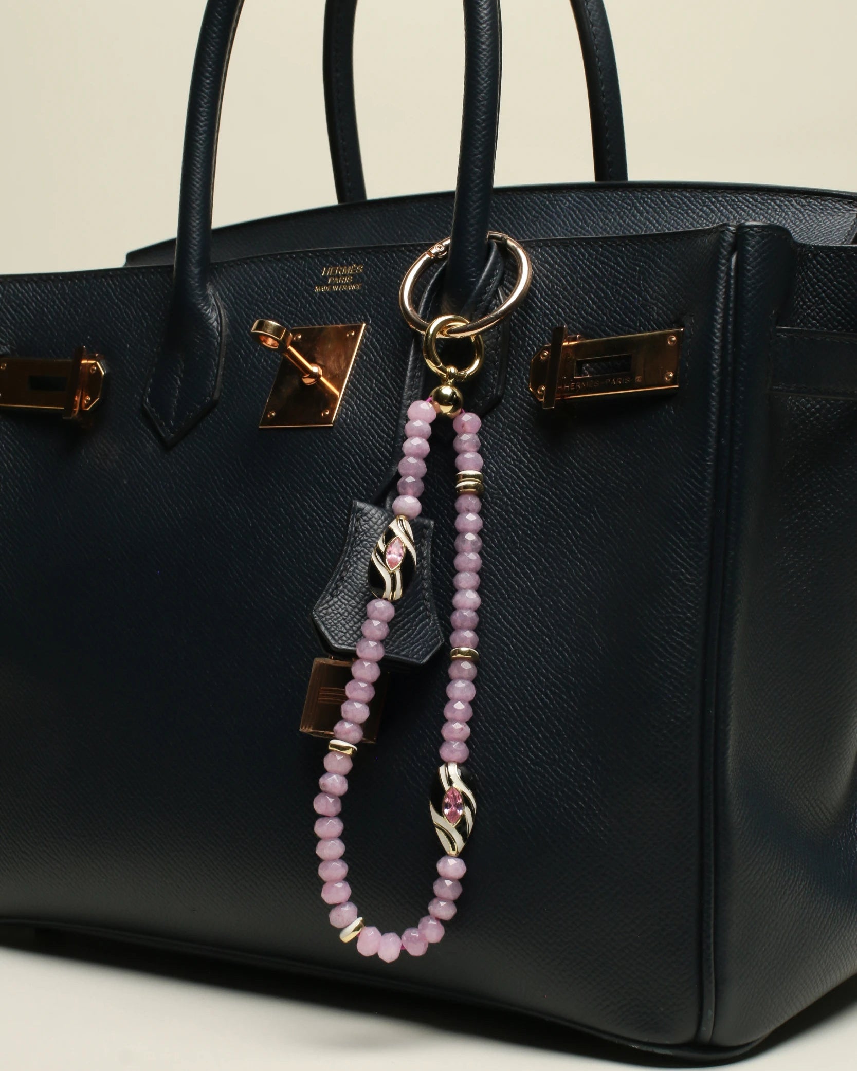 A structured black handbag adorned with The Minou Bag Charm in Lavender, highlighting its soft lavender quartz beads, gold-plated accents, and striking zebra-striped Evil Eye charm for a luxe finish.