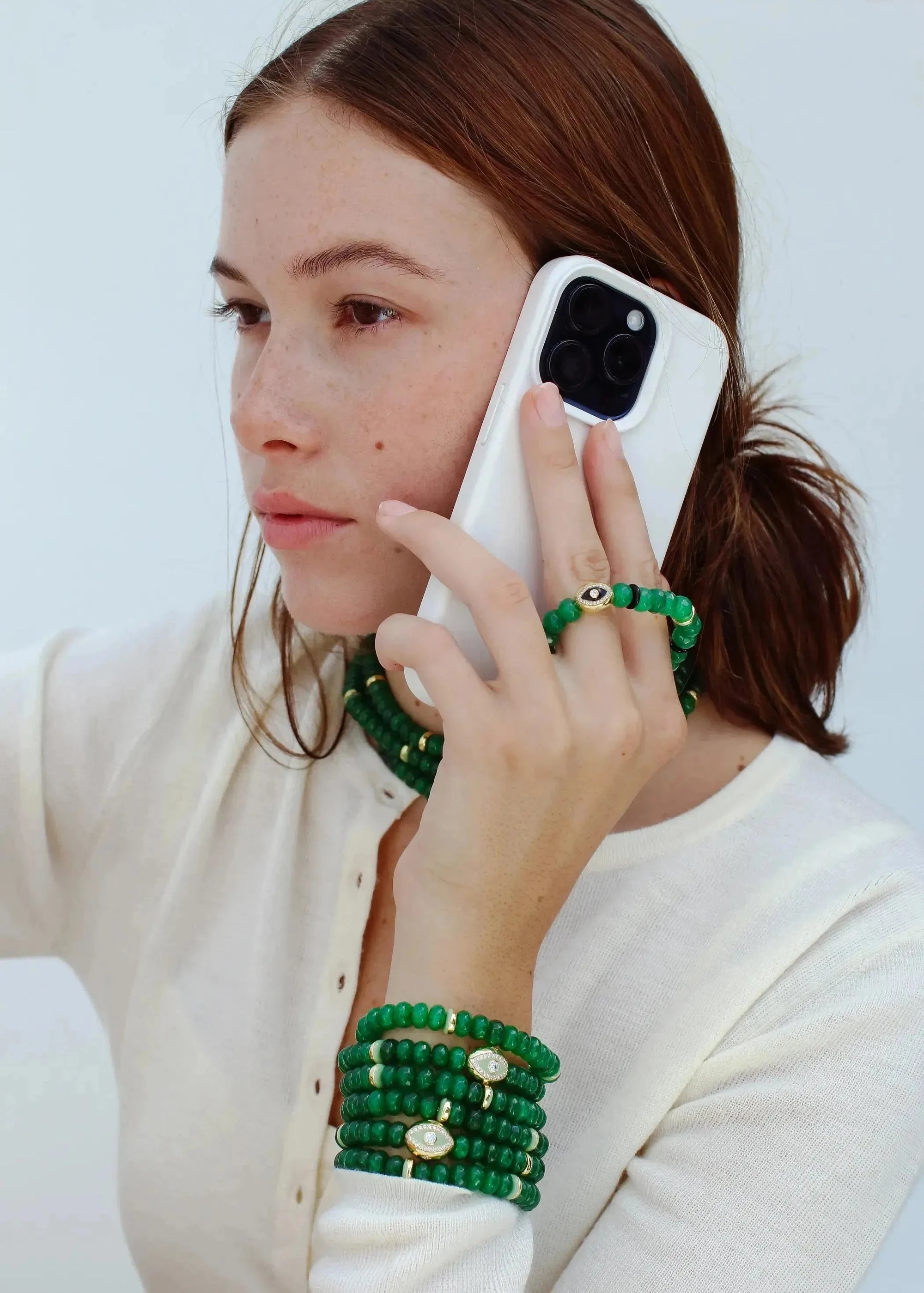 Model holding a phone with one hand, wearing the Leah Mini Wristlet in emerald as a phone accessory. The matching Leah bracelet set on the wrist and Lilou Necklace adds a layered, stylish touch to the look.