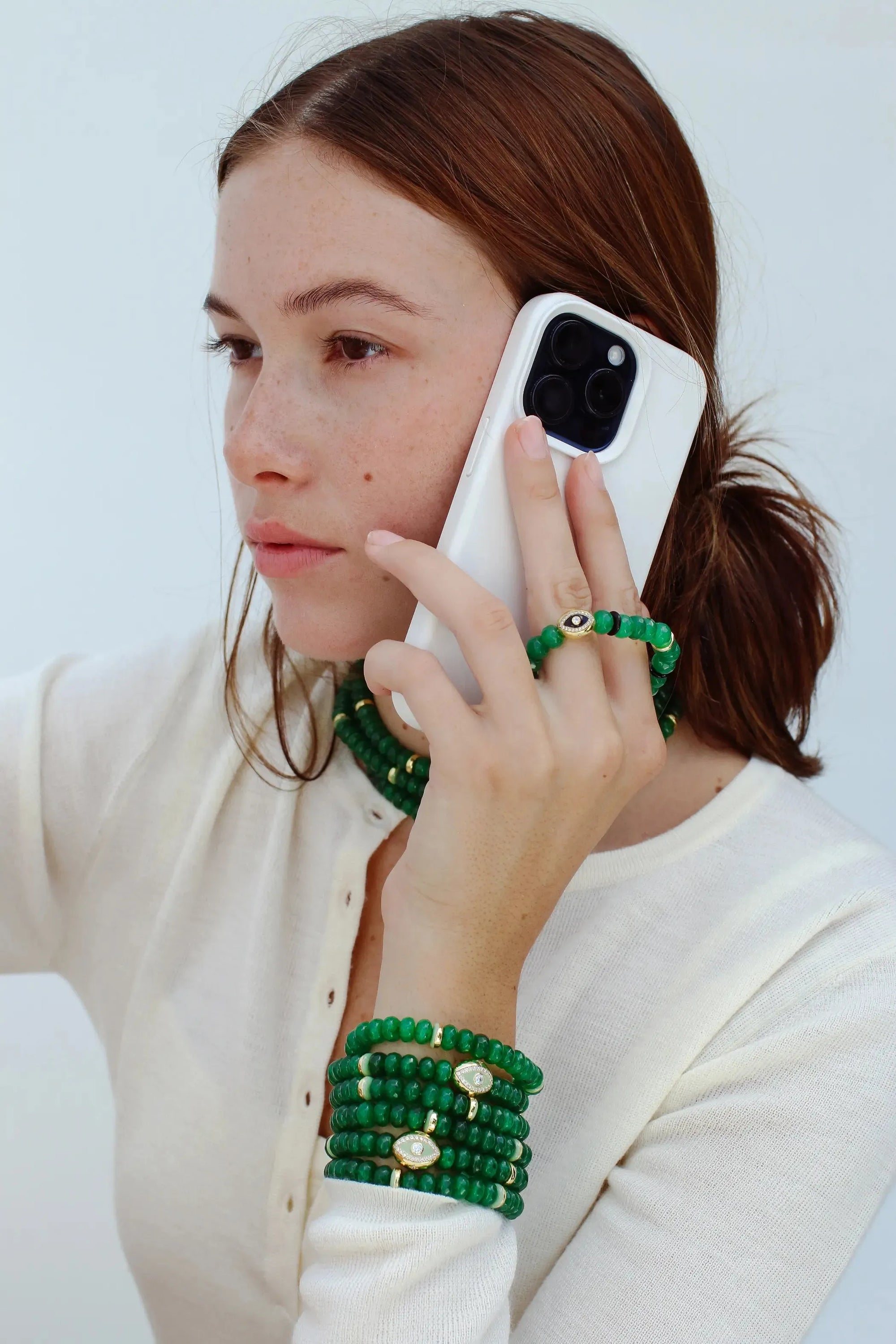 Model holding a phone with one hand, wearing the Leah Mini Wristlet in emerald as a phone accessory. The matching Leah bracelet set on the wrist and Lilou Necklace adds a layered, stylish touch to the look.