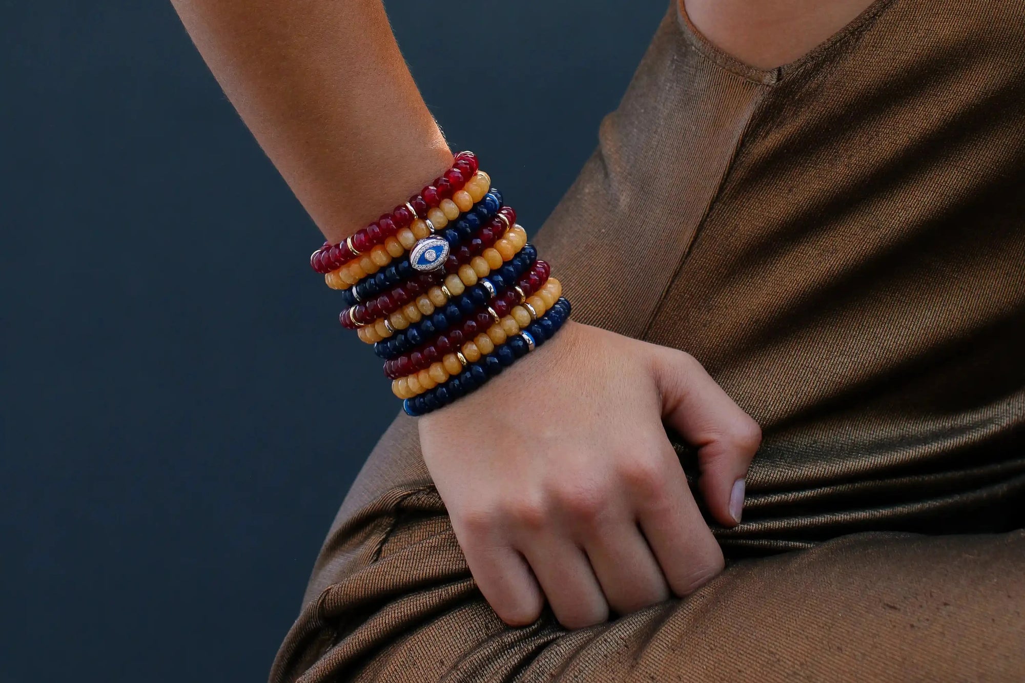 A stylish stack of Zoe Tangerine, Zoe Cherry, and Leah Sapphire bracelets elegantly wraps around a woman's wrist, paired with a brown outfit, showcasing vibrant colors and texture combinations.