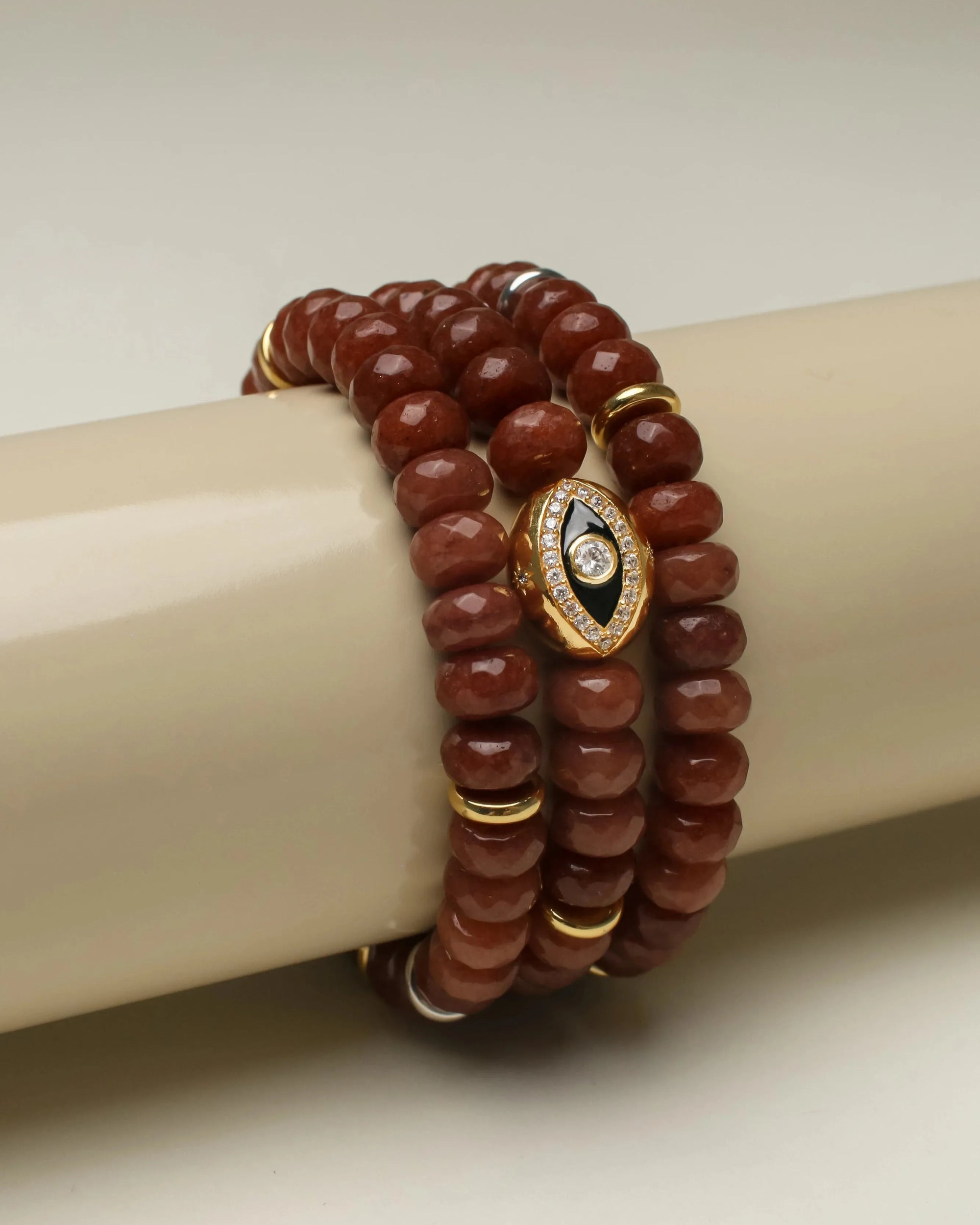 Stacked Leah bracelet set in caramel, showcasing polished faceted beads accented with gold details and a bold gold evil eye charm featuring a black center and crystal embellishments, elegantly wrapped around a neutral display.