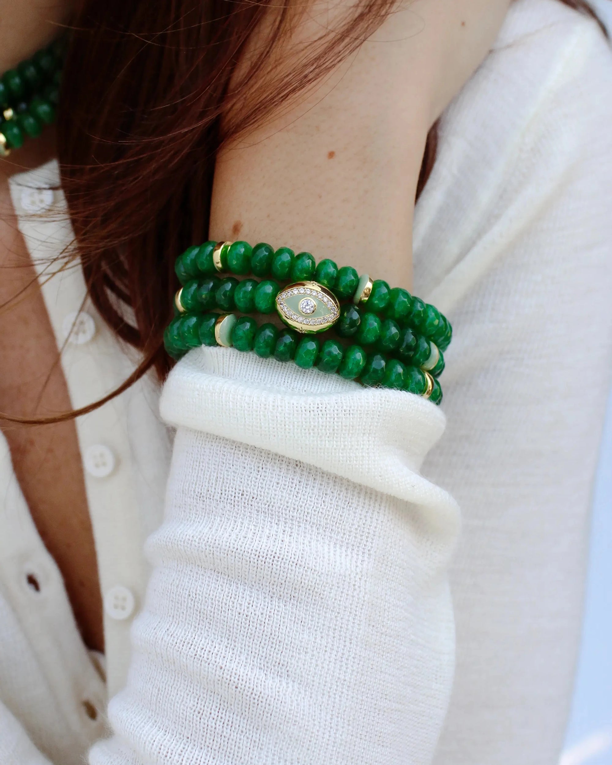 Close-up view of a woman's arm wearing a stack of emerald green Leah bracelets adorned with a central eye charm and gold accents, styled with a white knit top for a chic, elegant look.