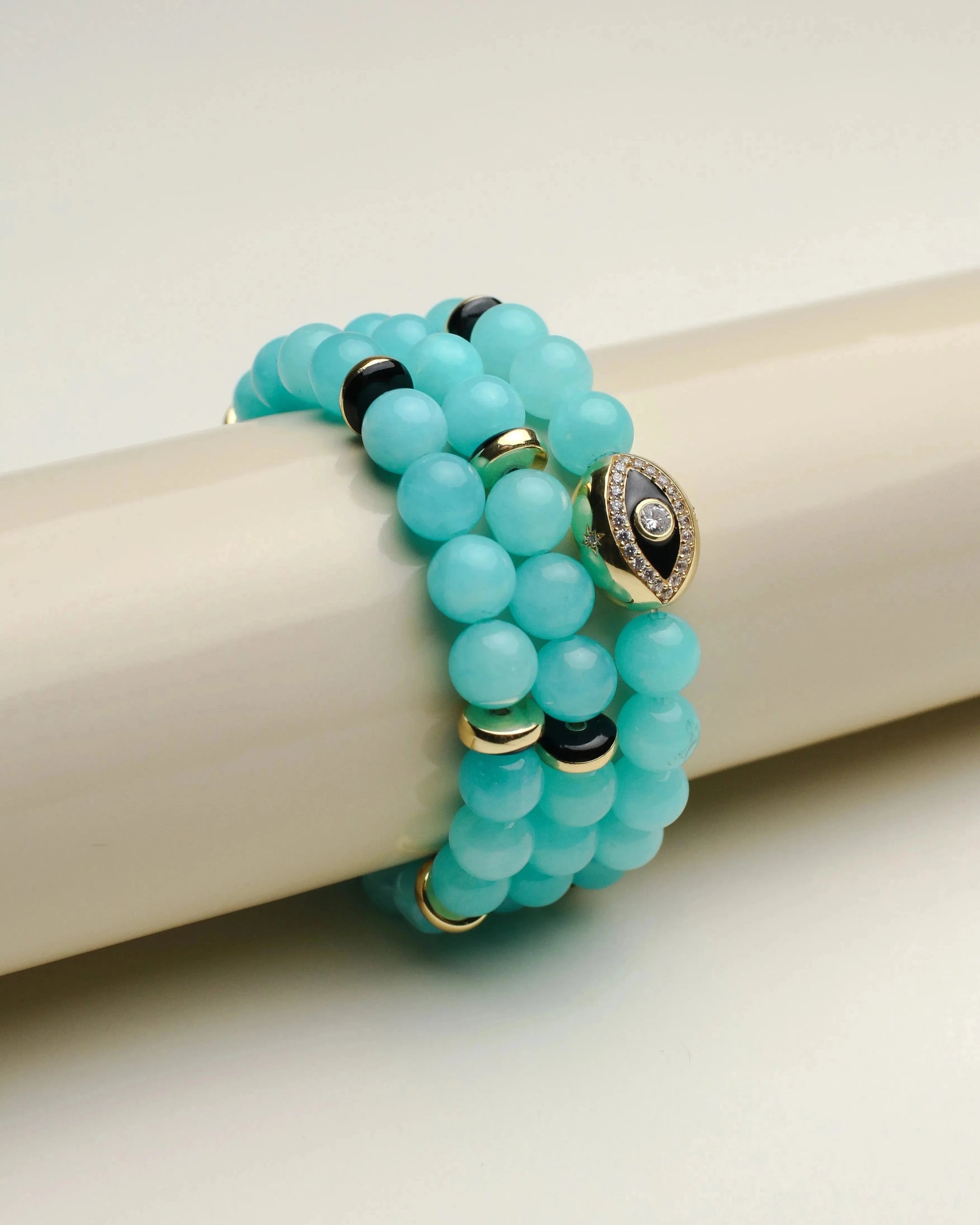 Leah aqua bracelet set of three displayed on a smooth cylindrical stand, showcasing the bright blue beads and gold embellishments with a gold evil eye charm.