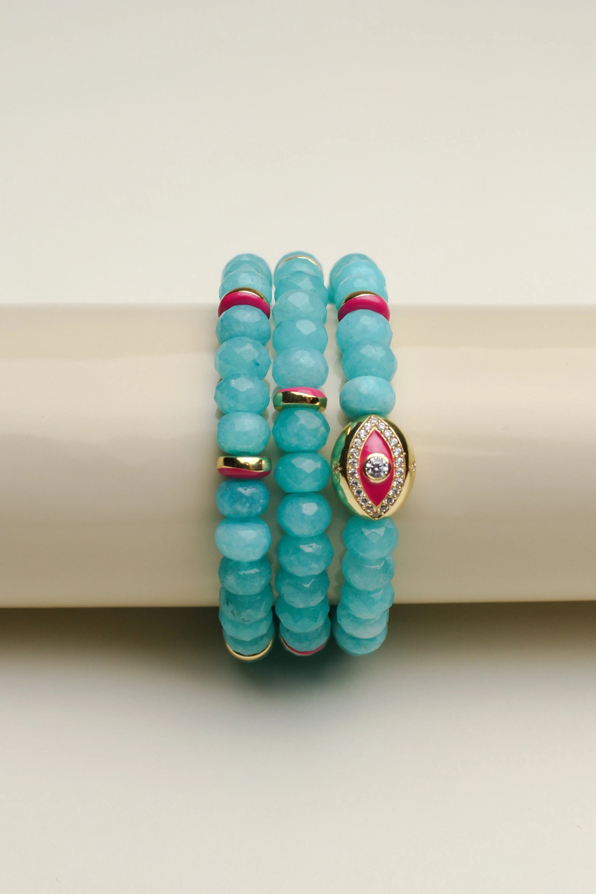 The Leah turquoise bracelet set displayed on a cylindrical stand, showcasing the bright blue-green beads with pink and gold detailing and an eye-catching central eye charm.