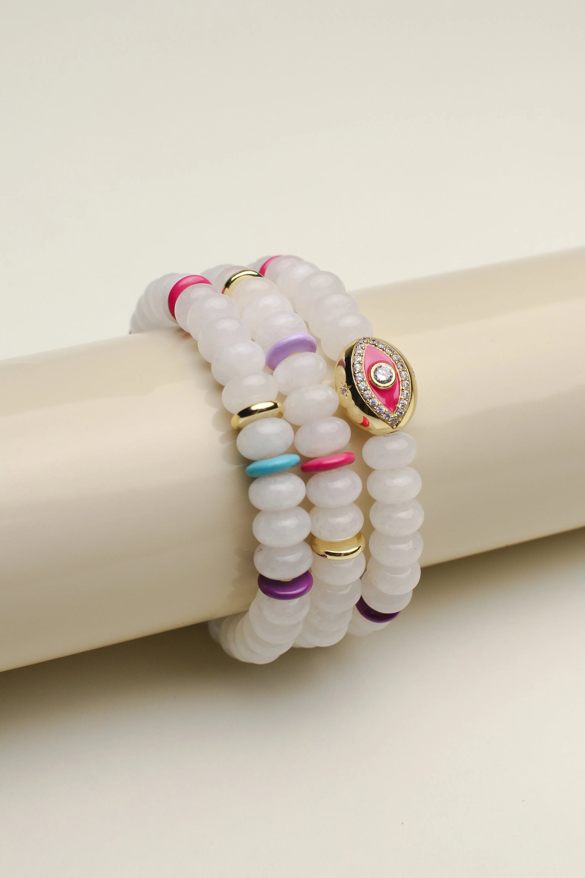 Leah white bracelet set of three displayed on a cylindrical stand, showcasing the smooth white beads with vibrant pink, blue, and gold accents and a central eye charm.