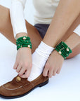 Model seated, wearing multiple stacks of the Leah Emerald Bracelet set on both wrists. The vibrant green quartz beads, accented with gold dividers and eye charms with CZ diamonds, complement the casual yet stylish outfit of white socks, brown loafers, and a cream top. The look blends effortless elegance with playful layering.