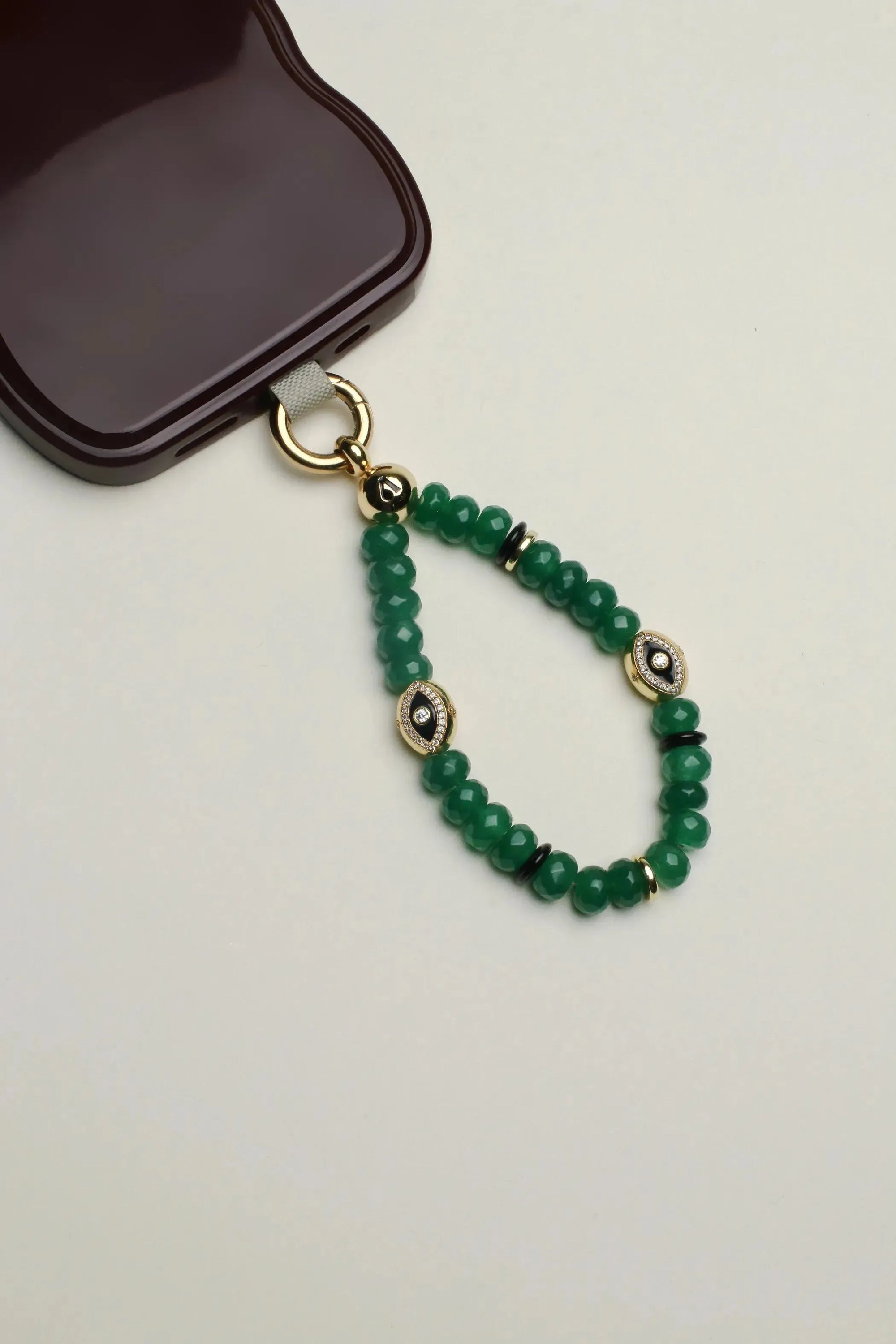 Close-up of the Leah mini Wristlet in emerald quartz attached to a sleek maroon phone case. The design features vibrant green beads, gold-plated accents, and an eye charm, blending elegance with practicality.