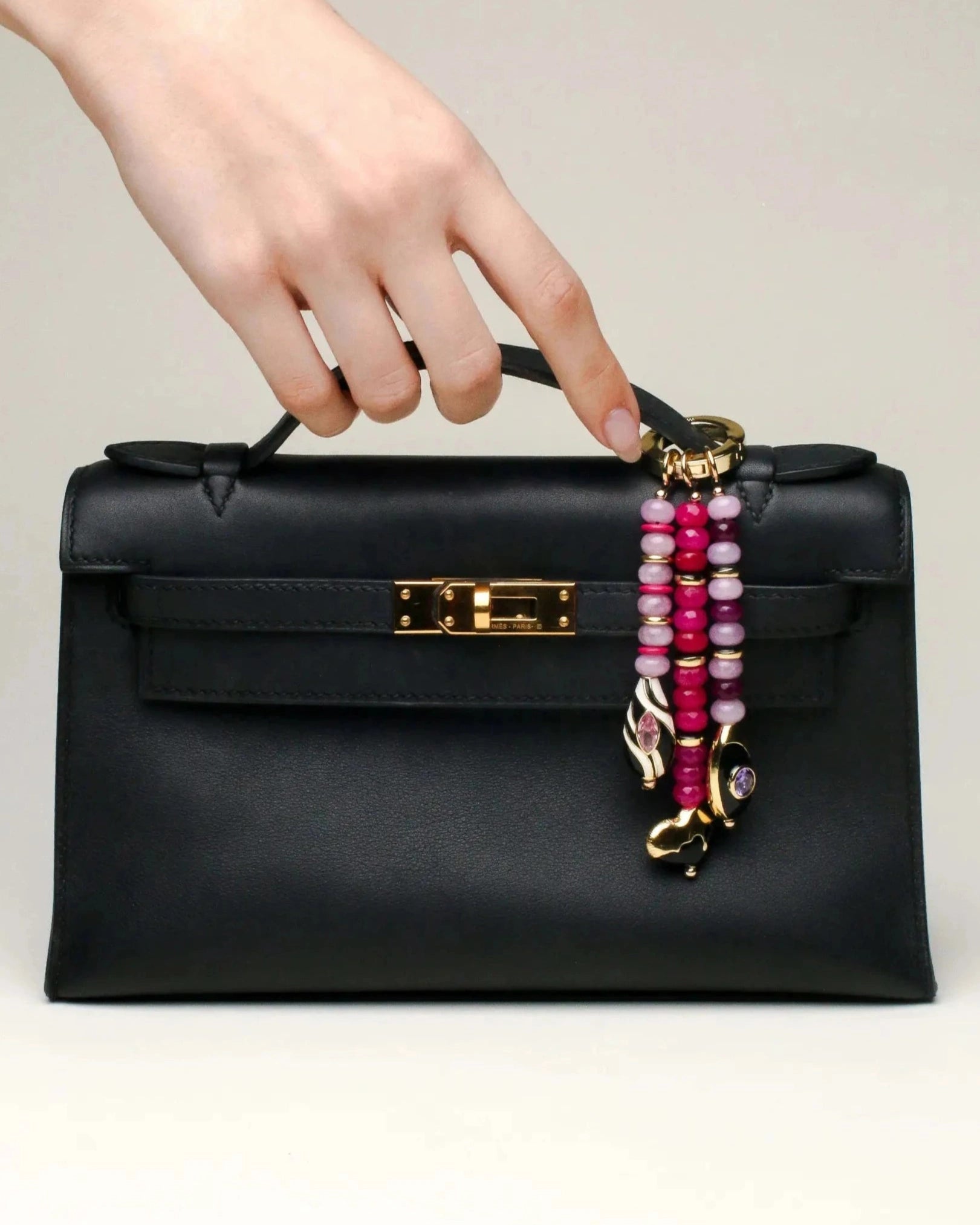 Close-up of a black handbag accessorized with the Liora charm. The three beaded strands in various shades of pink hang elegantly, each finished with gold-plated charms and gemstones.