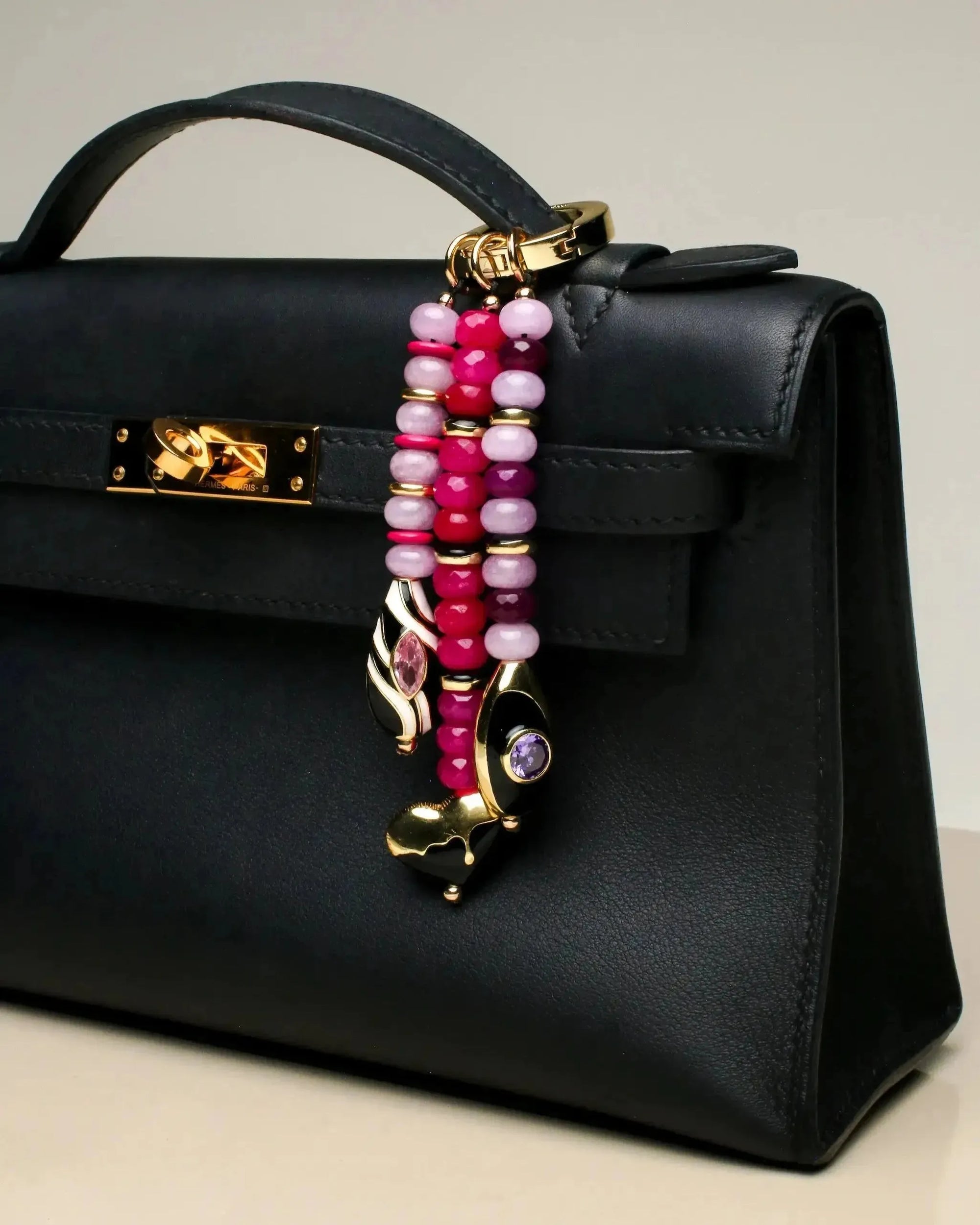 Close-up of a black handbag accessorized with the Liora charm. The three beaded strands in various shades of pink hang elegantly, each finished with gold-plated charms and gemstones.