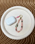 Mini Minou Rose Quartz Phone Charm placed on a cream round tray alongside accessories, attached to apple airpods and styled as a decorative flat lay to emphasize its luxury detail.