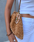 A tan woven suede bag with thin brown straps, styled with a pink Mini Minou charm made of rose quartz beads and gold accents, clipped to the bag. The model wears a white crop tank, white jeans, and a brown beaded bracelet.