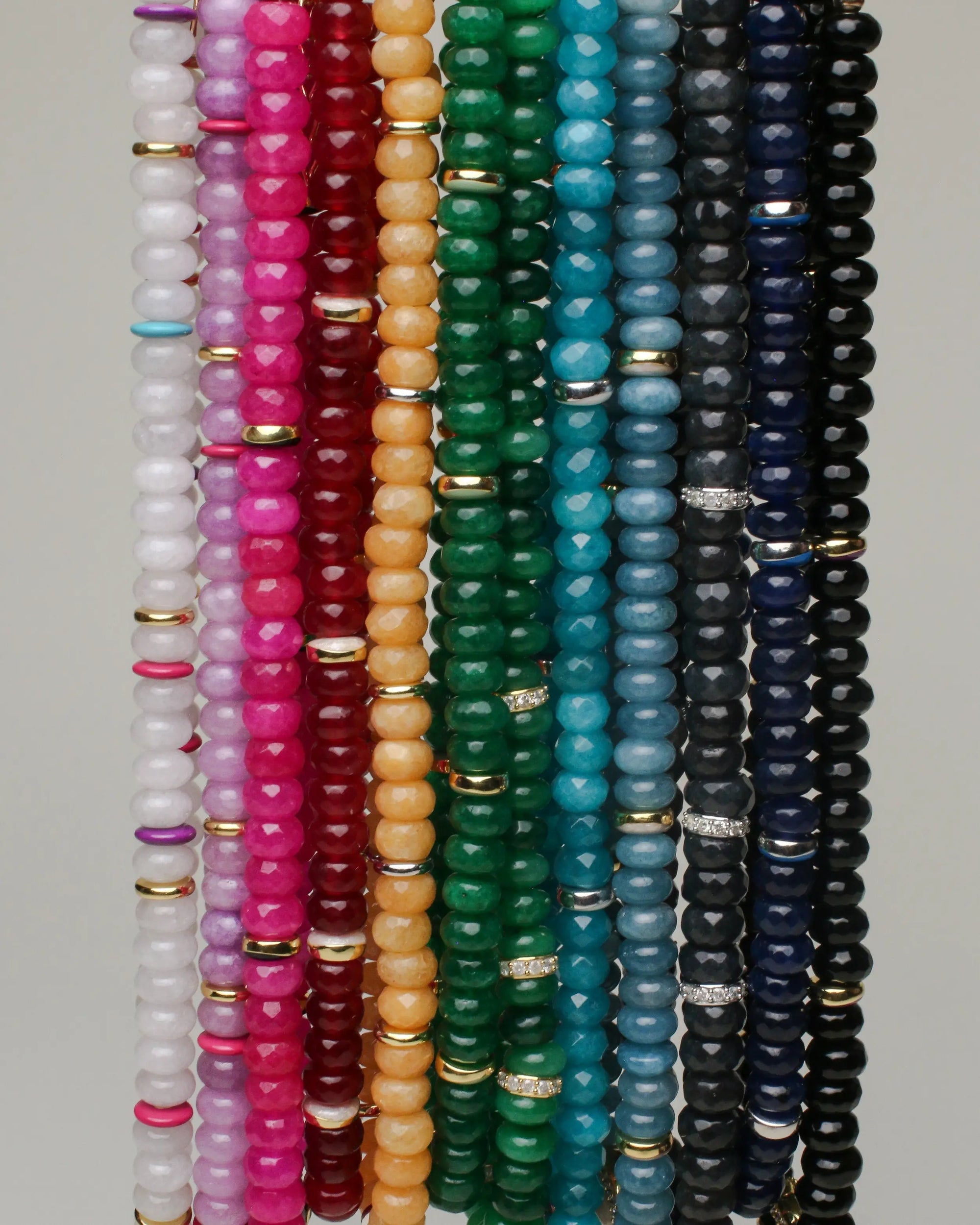 A vibrant display of stacked beaded necklaces showcasing a range of gemstone colors, including white, pink, red, orange, green, turquoise, blue, and black beads with gold dividers, highlighting the intricate craftsmanship and color diversity of each necklace."