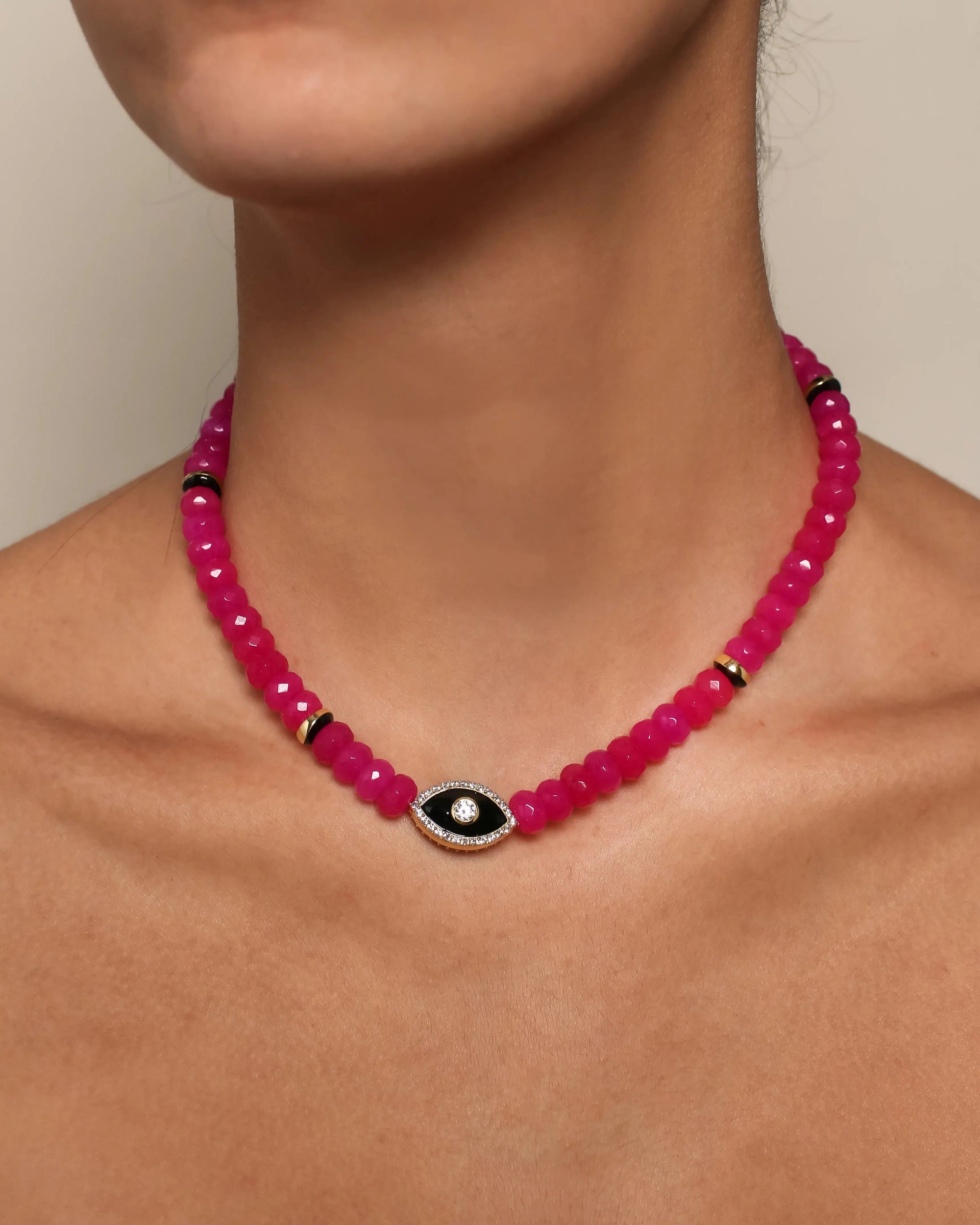 Close-up of a vibrant pink beaded Stella necklace featuring a striking black and gold evil eye charm with a central crystal accent, worn around the neck.