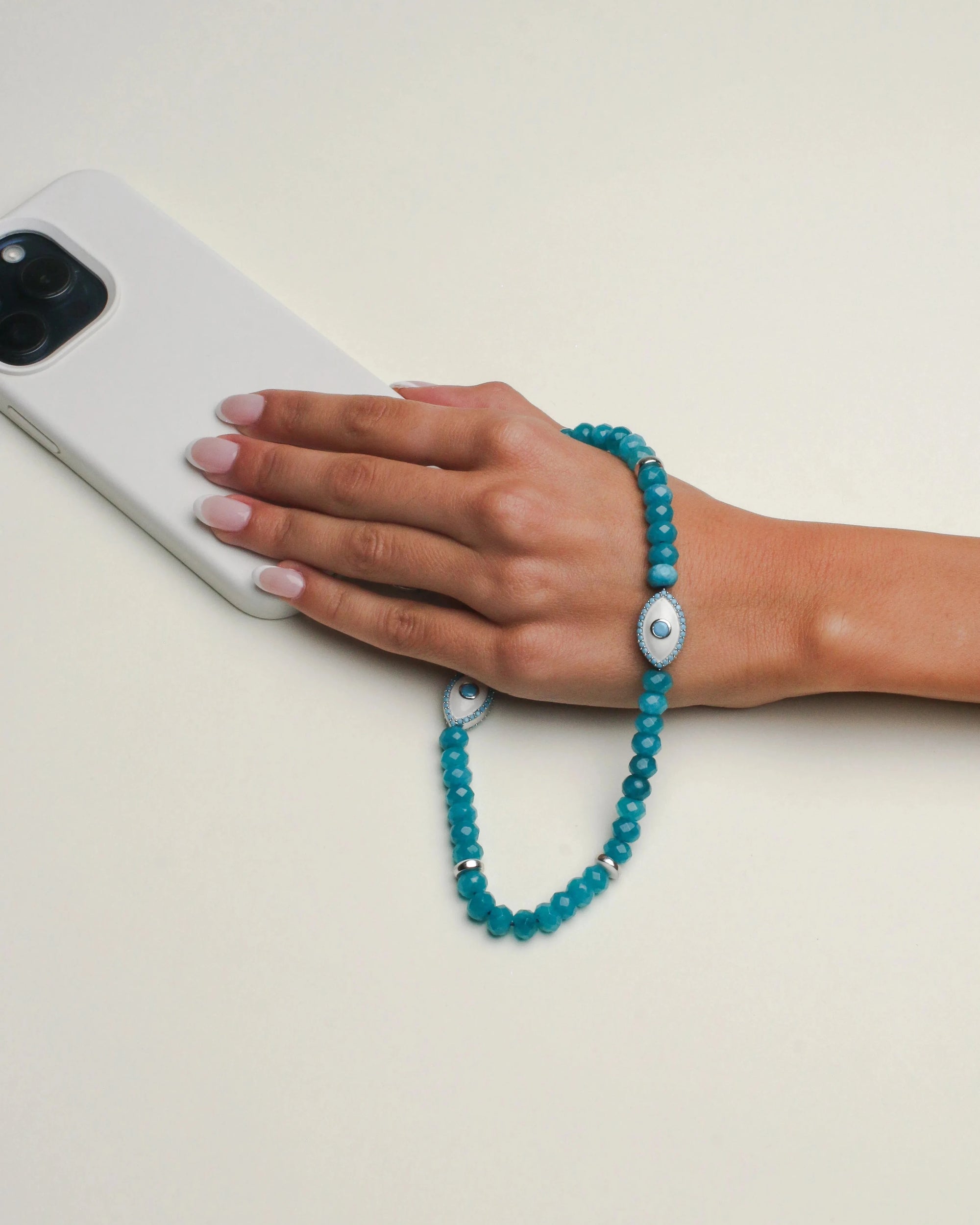 Hand holding a phone accessorized with the Stella wristlet in vibrant turquoise quartz and evil eye charm, highlighting its protective and fashionable design.