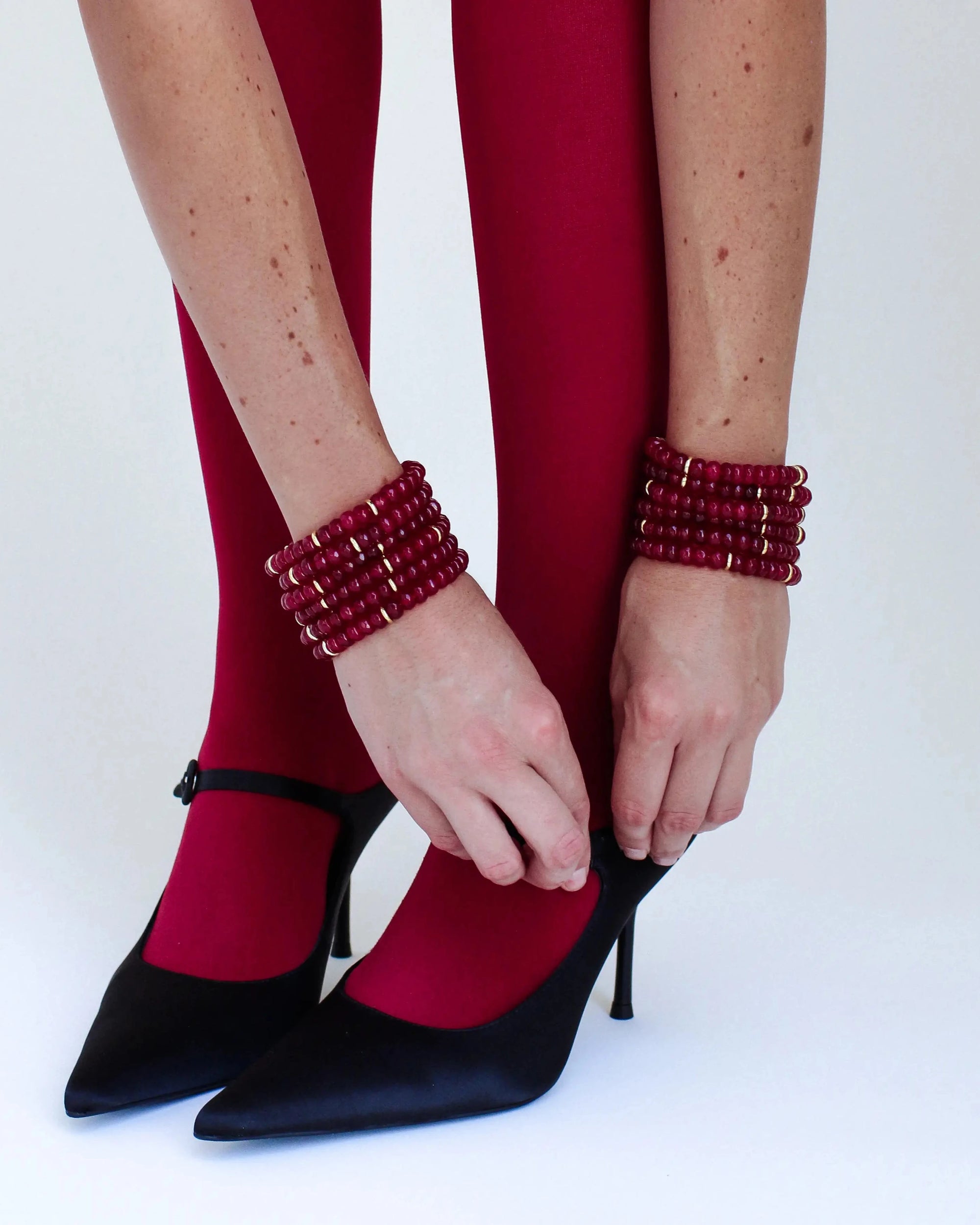 Close-up of Zoe Cherry bracelets worn around the wrists of a model wearing red tights and black heels, elegantly tying her shoe and highlighting the bold red tones of the accessories.