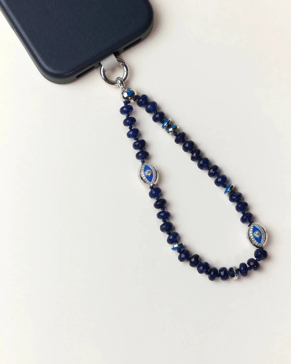 Flat lay of the Sapphire Leah Phone Charm attached to a black phone, showcasing deep-blue gemstone beads and rhodium-dipped evil eye accents.