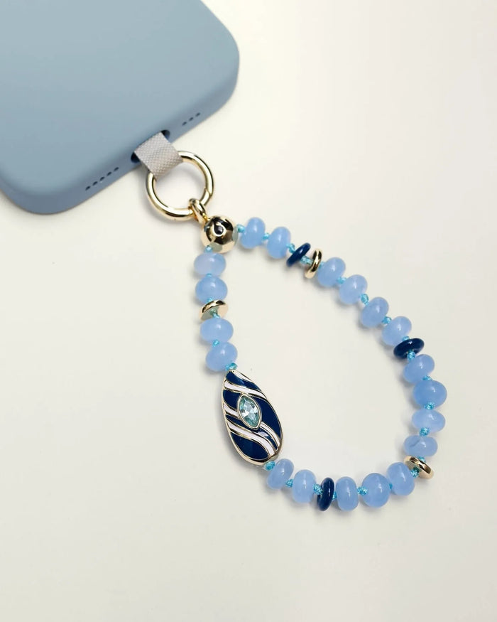 Mini Minou Aquamarine Quartz Phone Charm attached to a light blue phone case, featuring aqua-toned beads with a gold zebra evil eye charm.